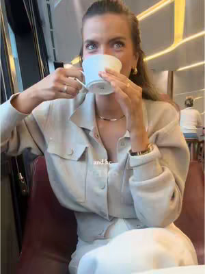 A woman is holding a white cup to her mouth, appearing to be drinking. The background is blurred, suggesting a public space like a restaurant or train. She is wearing a beige jacket and simple jewelry.