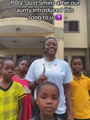 A group of children and a woman are standing in front of a building. The woman is in the center, pointing her index finger up, while the children surround her. The text overlay is present at the top of the image.