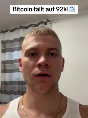 A man with short blonde hair and a slight beard stands in front of a blurred background of window curtains. He wears a white tank top and a gold chain. A text overlay at the top reads, 'Bitcoin fällt auf 92k!' with a downward arrow symbol indicating a price drop.