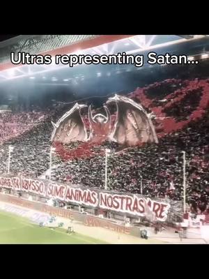 The image captures a crowded stadium with a large display created by the fans. The display appears to be a stylized image of a demonic creature, possibly a dragon or a similar mythological figure. The dominant colors are red, black, and grey, suggesting a dark or intense theme. Below this central image, a banner with text in a foreign language is also visible. The overall impression is one of grandeur, intensity, and possible religious or ideological symbolism.