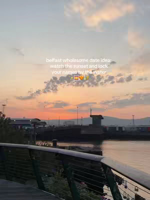 A serene sunset over a body of water, with a bridge and silhouetted structures in the foreground. Wispy clouds fill the sky, and the overall mood is peaceful. Text overlayed suggests a date spot in Belfast.