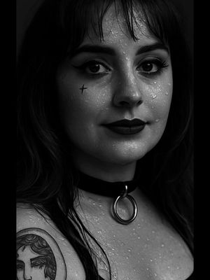 A black and white close-up portrait of a woman with dark hair, a cross tattoo, and a black choker. She has droplets of water on her face. Her lips are dark and her eyes are looking at the camera.