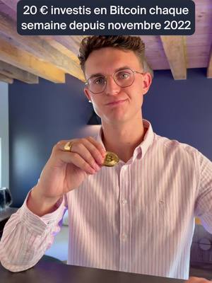 A man wearing glasses and a striped shirt holds a gold Bitcoin coin, smiling at the camera. Text overlay reads '20 € investis en Bitcoin chaque semaine depuis novembre 2022.' The background includes a dark wall and part of a ceiling with wooden beams.