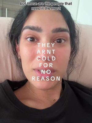 A close-up shot of a woman's face. The text overlay reads "THEY ARNT COLD FOR NO REASON." She appears to be looking directly at the camera with a serious expression. The background is a light-colored, blurred setting, possibly indicating a bedroom or indoor space. She is wearing a black top and a delicate gold necklace. The overall composition focuses on her face and the text overlay.