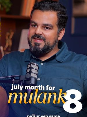 A medium shot of a man with a beard, wearing a dark blue collared shirt, sitting in front of a microphone. Text overlays in yellow and white read, 'july month for mulank 8' and 'pe aur yeh same'. The background appears to be a dark room with a bookshelf and other indistinct objects.