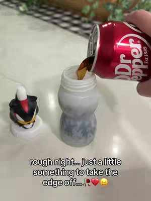 A person is pouring Dr. Pepper soda into a white, Shadow the Hedgehog themed cup. In front of the cup is a Shadow figurine with a black and red color scheme. The scene is set on a white surface with a blurred background.