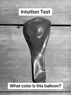 A black and white image featuring a deflated balloon against a wooden background. The balloon is positioned vertically in the frame, taking up most of the space. Above the balloon, there's a rectangular text overlay that says 'Intuition Test'. Below the balloon, another text overlay reads 'What color is this balloon?'. The wood has a grain and the image has a grainy texture.