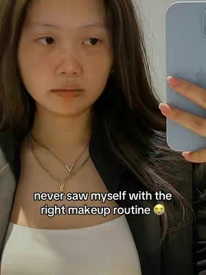 A selfie-style shot featuring a young woman looking slightly off-camera. She is holding a light blue phone, partially visible, towards the right side of the frame, while her own face takes up most of the space. She's wearing a white top and a black blazer. The background is a light-colored wall or surface, suggesting an indoor setting. The text overlay reads: "never saw myself with the right makeup routine 🥺".