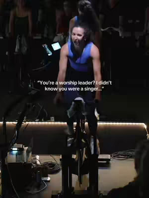 A vertical video shows a woman on a stationary bike in a cycling studio. She appears to be working out, and there is text overlayed on the image. The image is dark with some lights in the background.