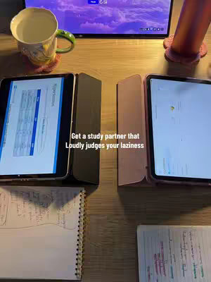 The image is a top-down shot of a desk with two open iPads placed side-by-side. Between them is a stack of what appears to be a closed black book or case. On the left is an iPad showing a spreadsheet; on the right, the screen is blank. Below, in the foreground, is a spiral notebook and the edge of another book, presumably for notes or studying. To the side, a partially visible mug and a laptop screen, along with a coaster, suggest a study environment. The overall color palette is neutral with warm lighting.