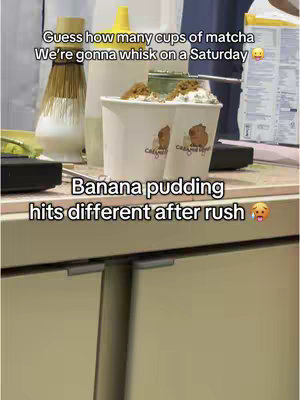 The image shows two cups of a dessert, potentially banana pudding, sitting on a counter. The cups are white with a logo. The banana pudding topping is visible. Behind the cups is a matcha whisk, containers, and other items. The scene is brightly lit, and the overall composition focuses on the food item.