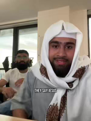 A selfie-style shot features two men in an indoor setting. The primary subject, positioned in the foreground, is a man wearing a white head covering and a grey garment, giving a slight smile. The secondary figure, blurred slightly in the background, also appears to be a male, wearing glasses and a white t-shirt. The background is minimal, featuring plain walls and a glimpse of a window or other structure.