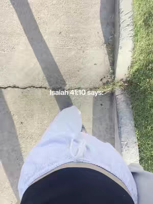 A first-person perspective shot walking on a paved surface. Below the frame focuses on the midsection, from the waist down, and includes part of what appears to be a white cap. The scene is lit with bright sunlight, creating a strong shadow on the pavement. To the right, there is grass and some greenery. Text overlay is positioned near the center, providing context related to a verse.