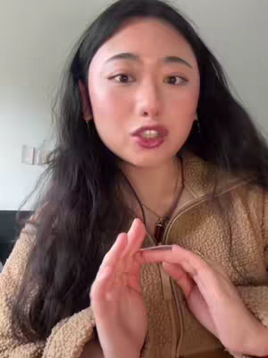 A woman with long dark hair is centered, talking and gesturing with her hands. She is wearing a brown fleece jacket. The background is a plain, off-white wall.