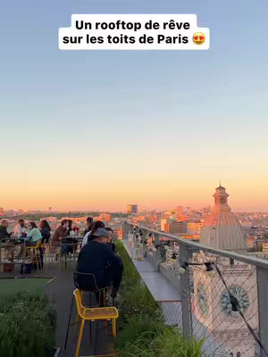 An eye-level shot of a rooftop bar at dusk. In the foreground, a group of people sit at tables, some on yellow stools, and a few leaning on a small platform, enjoying the view. The background reveals a cityscape with buildings and a dome, all under an orange and blue sunset sky.