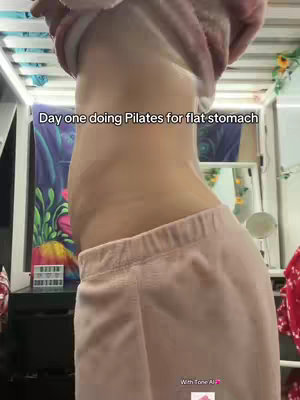 The image shows a woman from the side, focusing on her midriff and what appears to be her stomach area. She is wearing pink sweatpants and a top, with her arm raised and holding up her top to show her side profile, highlighting her waist. Text at the top reads 'Day one doing Pilates for flat stomach'. The background includes a glimpse of a bed frame, a curtain with colorful patterns, and a few decorative items.
