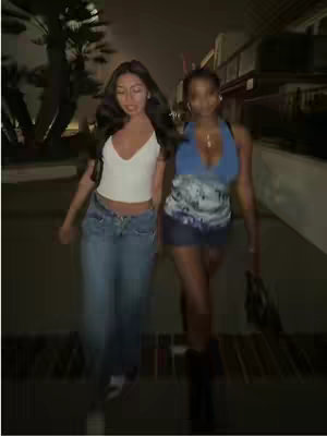Two young women walking side by side down a dark city street at night. The woman on the left is wearing a white top and jeans, while the woman on the right is wearing a blue and white patterned top and skirt. Both are smiling.