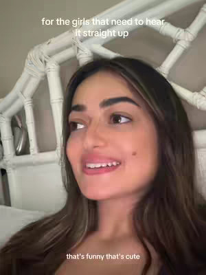 A woman with long, dark hair is shown smiling, her head and shoulders in frame. She is looking slightly off to the side, appearing to be speaking. Above her, text reads, 'for the girls that need to hear it straight up', and below, 'that's funny that's cute'. In the background is a white headboard.