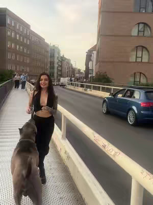 A woman with long dark hair is walking on a concrete walkway with a dog, the sky is overcast and the woman looks friendly, a street and buildings make up the rest of the background. A gray dog is in the foreground walking beside the woman.