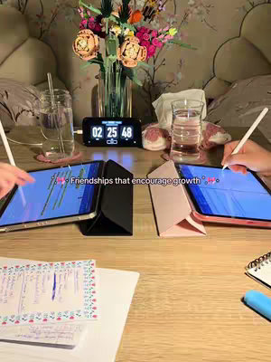 The image shows a close-up of a table with two tablets, two pairs of hands holding styluses. A clock sits in the background. Two glasses of water, some pink decorations. And a vase with flowers. The cover gives the impression that someone is studying or working together.