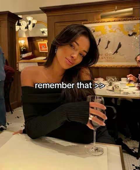 A woman with long, dark hair is seated at a restaurant table, holding a glass of red wine. She wears a black, off-the-shoulder top, and the background shows a restaurant setting.