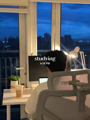 A wide shot of a room with a large window overlooking a city skyline at dusk. A person is sitting at a desk, facing away from the camera, and working on a computer. On the desk are a laptop, a drink, and a potted plant. The text overlayed on the image says "studying" and the time as "6:18 PM".