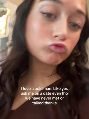 A close-up selfie of a young woman with dark hair and fair skin, making a kissing face. The background is blurred, suggesting an indoor setting.