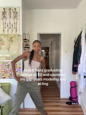 A young woman stands in a brightly lit hallway, striking a pose. She wears cargo pants and a tank top, with her hair pulled back. The background includes decorative wall art and a door leading to another room.