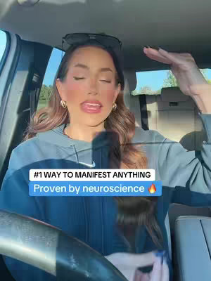 A woman is seated inside a car, facing forward with her eyes closed, and her hand raised, as if gesturing or making a point. The image has text overlay in the lower portion of the screen.