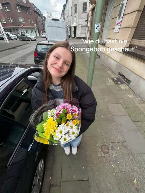 A young woman stands on a city street, smiling and holding a bouquet of colorful flowers. She is wearing a black jacket, a blue sweater, and jeans. The background shows a street with cars and buildings.