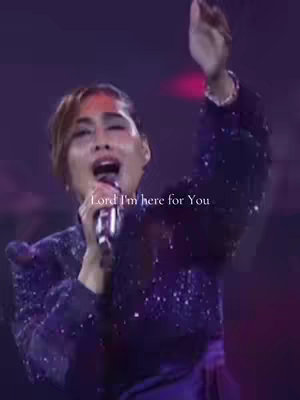 A woman with brown hair and a dark, sparkly top is singing into a microphone, her mouth open. She's holding the microphone with one hand and has her other hand raised up. The background is dimly lit, adding to a stage-like atmosphere. Text overlayed says, "Lord I'm here for You."