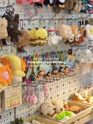 A brightly lit, vertical shot showcasing a shop display with merchandise prominently featured. The items include a variety of plush keychains, in the form of various animated characters, displayed on what looks like a grid of pegboard-style panels. The keychains are hanging on hooks, and some are arranged to show the variety of items. Below there are also some items in display within wooden shelves. The color palette is vibrant, with primary hues of white, yellow, brown, green, and orange. The overall composition is a tidy and inviting shop.