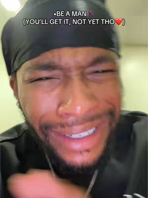 A close-up shot features a Black man, his face deeply expressive with a pained or strained look. He's wearing a black durag, and the lighting is such that it emphasizes his facial features. The background is blurred, keeping the focus on his face and the emotions he's conveying. Added text at the top suggests a message related to manhood.