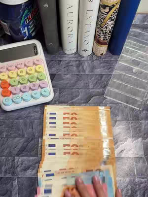 The image showcases a top-down view of financial organization. A colorful calculator with rainbow buttons is positioned on the left. Stacks of euro banknotes and a hand are present at the bottom. Behind these are several books and a jar of Nutella. The composition suggests a focus on budgeting or personal finance, with various props implying home-based financial activities.