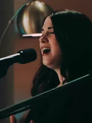 A medium shot shows a woman singing into a microphone with passion. She is positioned slightly off-center, with her face lit by warm light, eyes closed in devotion and her mouth open wide. A microphone stands prominently in the foreground. The background is blurred, suggesting a studio setting.