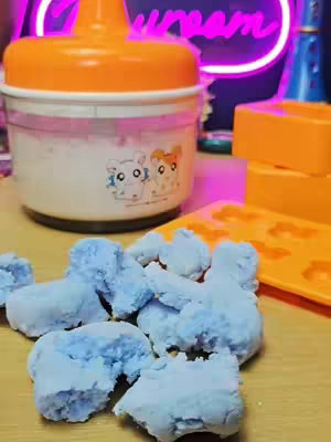 The image shows a close-up of a vintage Hamtaro Marshmallow Maker setup. An orange and white marshmallow maker filled with what appears to be white fluffy marshmallows is in the center-left. A pile of blue marshmallow shapes sits in the foreground. Behind the maker is an orange tray of molds with cloud shapes.