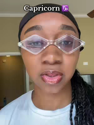 A close-up shot features a person with a dark head covering and diamond-shaped glasses. The person has visible facial features, including lips with lip gloss and a neutral expression. Overlaid at the top is the text "Capricorn" with a zodiac symbol. The background is blurred, suggesting an indoor setting.