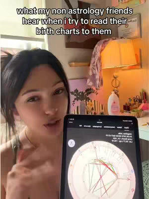 A young woman is shown in a selfie video, holding a tablet displaying a birth chart. She is looking at the camera with a slight frown. The background includes a shelf with items, a lamp, and a painting, creating a cozy indoor setting. The lighting appears to be warm.