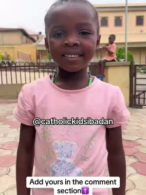 A young girl with a radiant smile stands in front of a building. She wears a light pink t-shirt with a teddy bear graphic and a beaded necklace. Text overlays appear at the bottom, inviting viewers to comment. In the background, another person sits on a structure behind her.