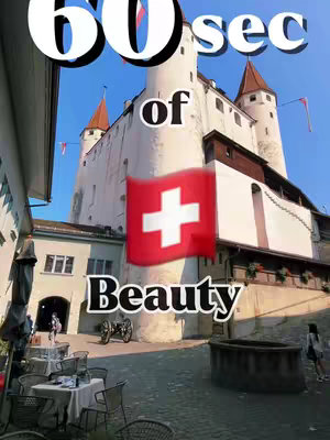 The cover image shows a classic European town square with a large castle in the background. A red and white Swiss flag is superimposed in front of the castle. The image has text added, promoting the location.