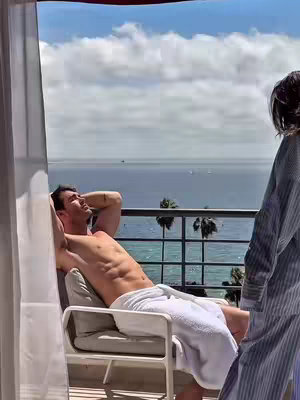 A man relaxes on a balcony chair overlooking a tranquil sea view. He is shirtless, wrapped in a white towel, and reclining with arms behind his head. Another person, partially visible, stands to the right in a striped robe. The sky is bright with clouds.