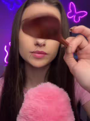 The image shows a young woman looking at the camera. She is holding a brown object in front of her face that partially obscures her eyes. She has long brown hair and is wearing a pink top. In the background are purple neon signs and a pink fluffy object is in the foreground.