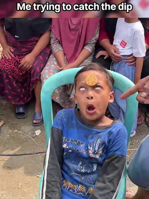 A young boy with a cookie placed on his forehead, eyes rolled upwards, mouth open in a surprised expression. In the background, several people are sitting or standing, some with their hands visible. The scene evokes a sense of anticipation or surprise.