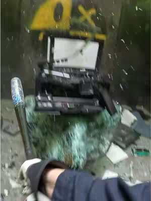 The image shows a person's arm holding a baseball bat about to smash a laptop on a surface covered in shattered glass. In the background, some painted yellow and black details can be seen on a hard object. The scene is action oriented.