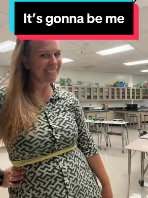 A woman with long blonde hair is smiling at the camera. She is wearing a black and white geometric patterned dress and a yellow measuring tape is wrapped loosely around it. The background suggests a classroom setting with whiteboards, cabinets, and lab equipment.