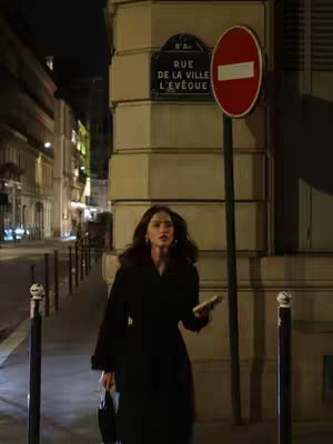 A woman walks down a Paris street at night, holding a small item. The scene is lit by streetlights and shop windows.