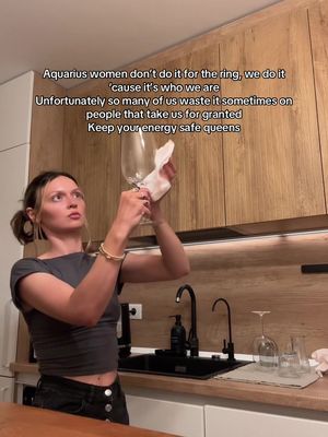 A woman in a kitchen setting, holding a wine glass and wiping it with a cloth. She is dressed in a casual outfit and is looking up and slightly to the left, seemingly focused on cleaning the glass. The background includes wooden cabinets and a modern kitchen design.