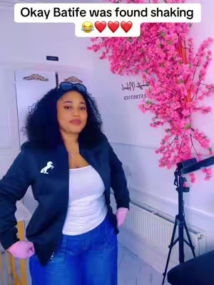 A woman with dark curly hair is centered in the frame, wearing a black jacket, a white top, and blue jeans. She has pink gloves on her hands and sunglasses atop her head. Behind her is a pink floral arrangement and a white wall.
