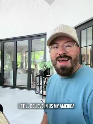 A man wearing glasses and a baseball cap smiles and speaks to the camera. The background includes a white wall and large glass doors leading outside.