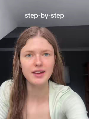 A young woman with long, light brown hair and fair skin is looking directly at the camera. She is wearing a light green top. Above her head is white text on a black background that reads "step-by-step". The background is somewhat blurred and shows elements of a room, including a window and wall.